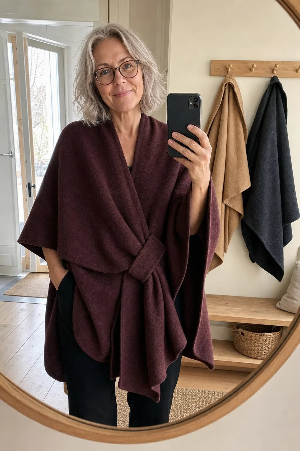 Kunde i vinrød Lovelle Poncho foran gangspeilet, med en kamel og en mørkegrå på knaggene ved siden av